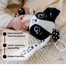 Load image into Gallery viewer, Black and white plush toy held by a child's hand on a soft surface, with text highlighting features.
