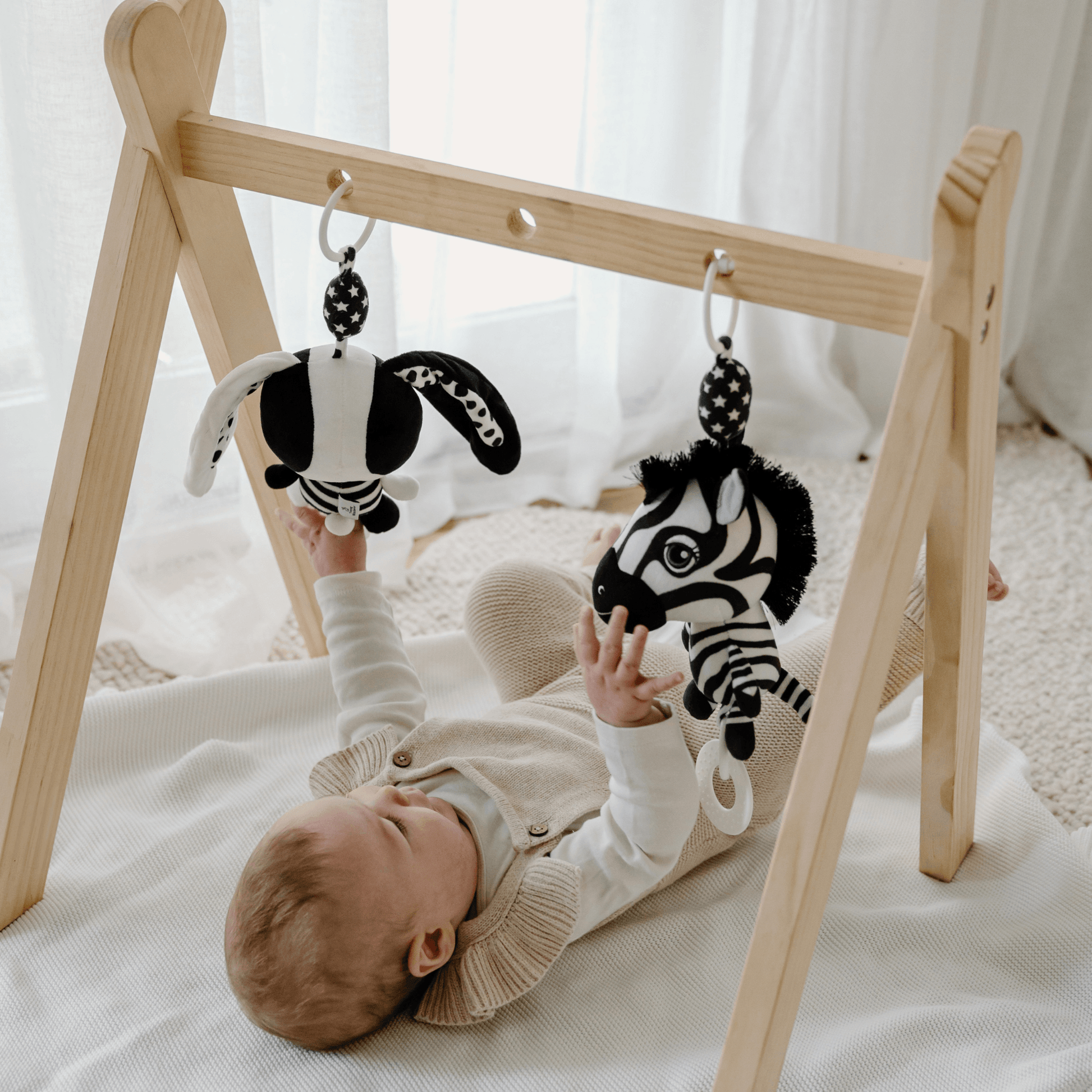 Baby reaching up to play with Ziggy the Zebra and a matching black and white Billie the Bunny toy hanging from a wooden baby gym 